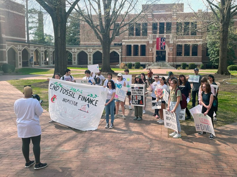 A group of people surrounded by academic buildings hold signs encouraging the University of Richmond to divest from fossil fuels and listen to a speaker holding a megaphone.