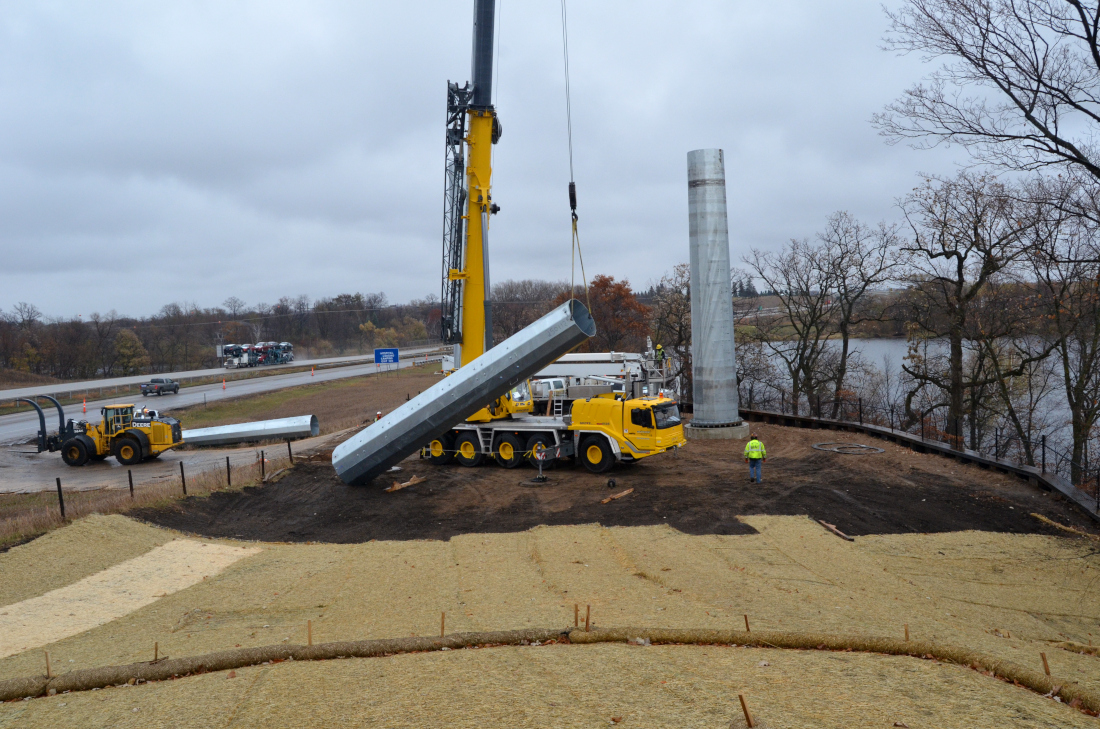 A heavy construction crane lifts a segment of a transmission tower into place along a rural expressway.